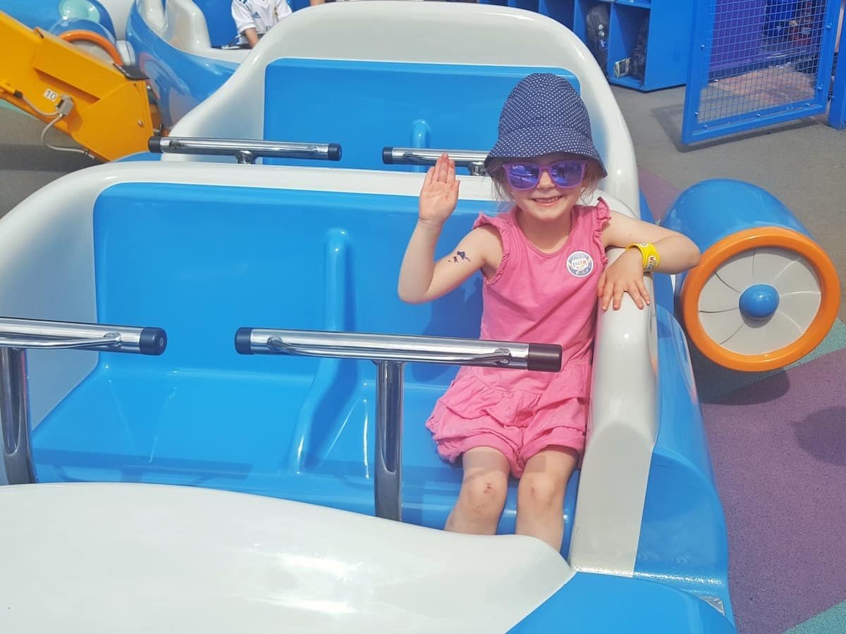 A little girl in a pink dress and dark blue spotted sun hat (Libby) enjoying a ride at Alton Towers smiling and waving to the camera on a plane with blue seats. 