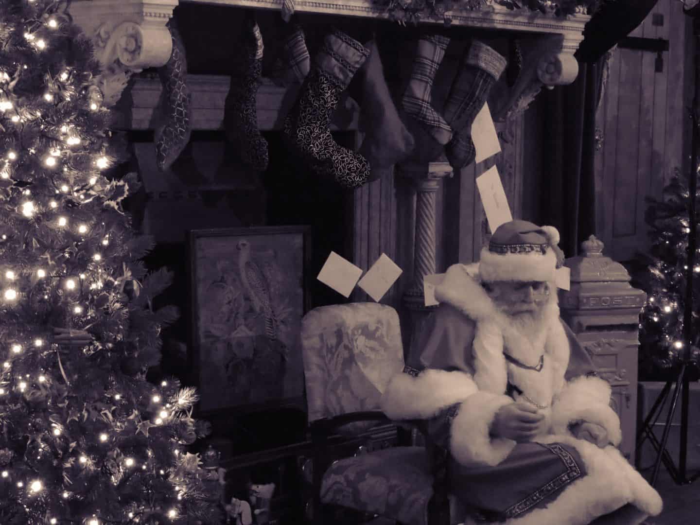 Santa sitting in front of a large fire place by a Christmas tree. Image is in black and white