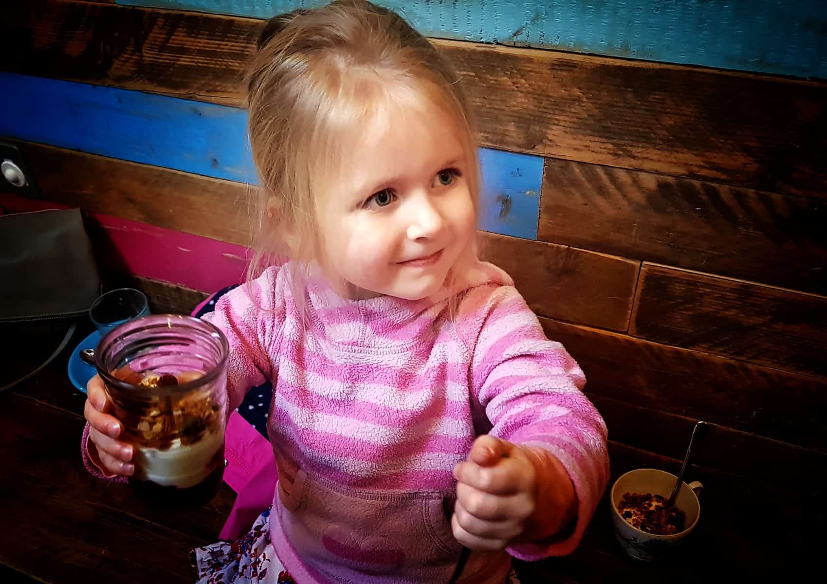 little girl in a pink jumper holding a pot of yoghurt and granola