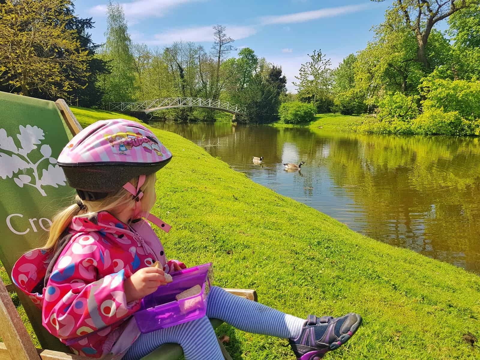Little girl wearing a cycling helmet and eating from a lunchbox, staring into the distance at a river with bridge over