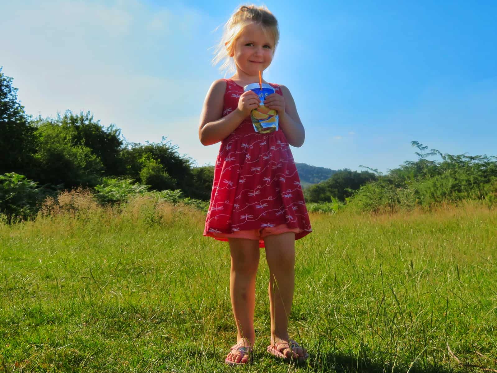 Little girl stood on grass drinking Capri-Sun with blue sky behind