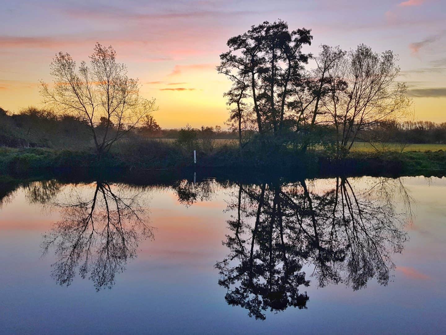 Outdoor swimming spot in Fladbury, Worcestershire | depicts the river at sunrise with reflections of the trees and sunrise colours of pink and yellow. 