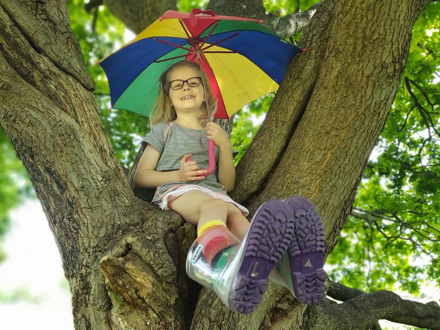 Little girl with rainbow umbrella sitting in a tree wearing squelch wellies and rainbow socks facing forwards