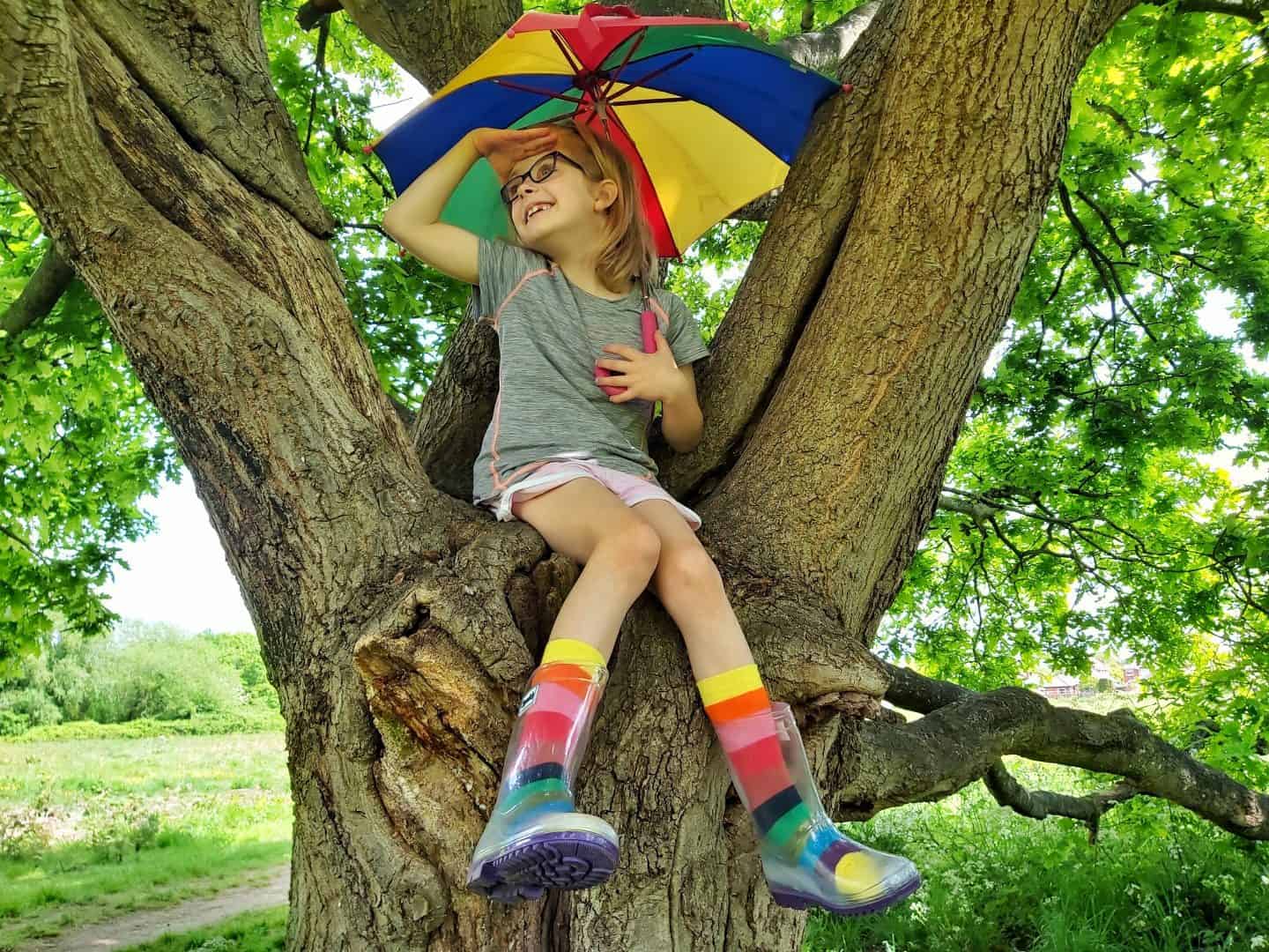 Little girl with rainbow umbrella sitting in a tree wearing squelch wellies and rainbow socks looking into the distance