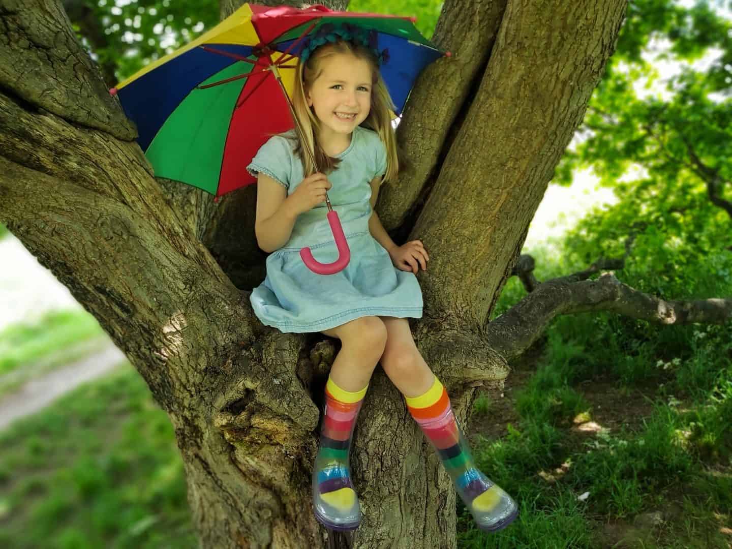 Little girl with rainbow umbrella sitting in a tree smiling and wearing squelch wellies and rainbow socks