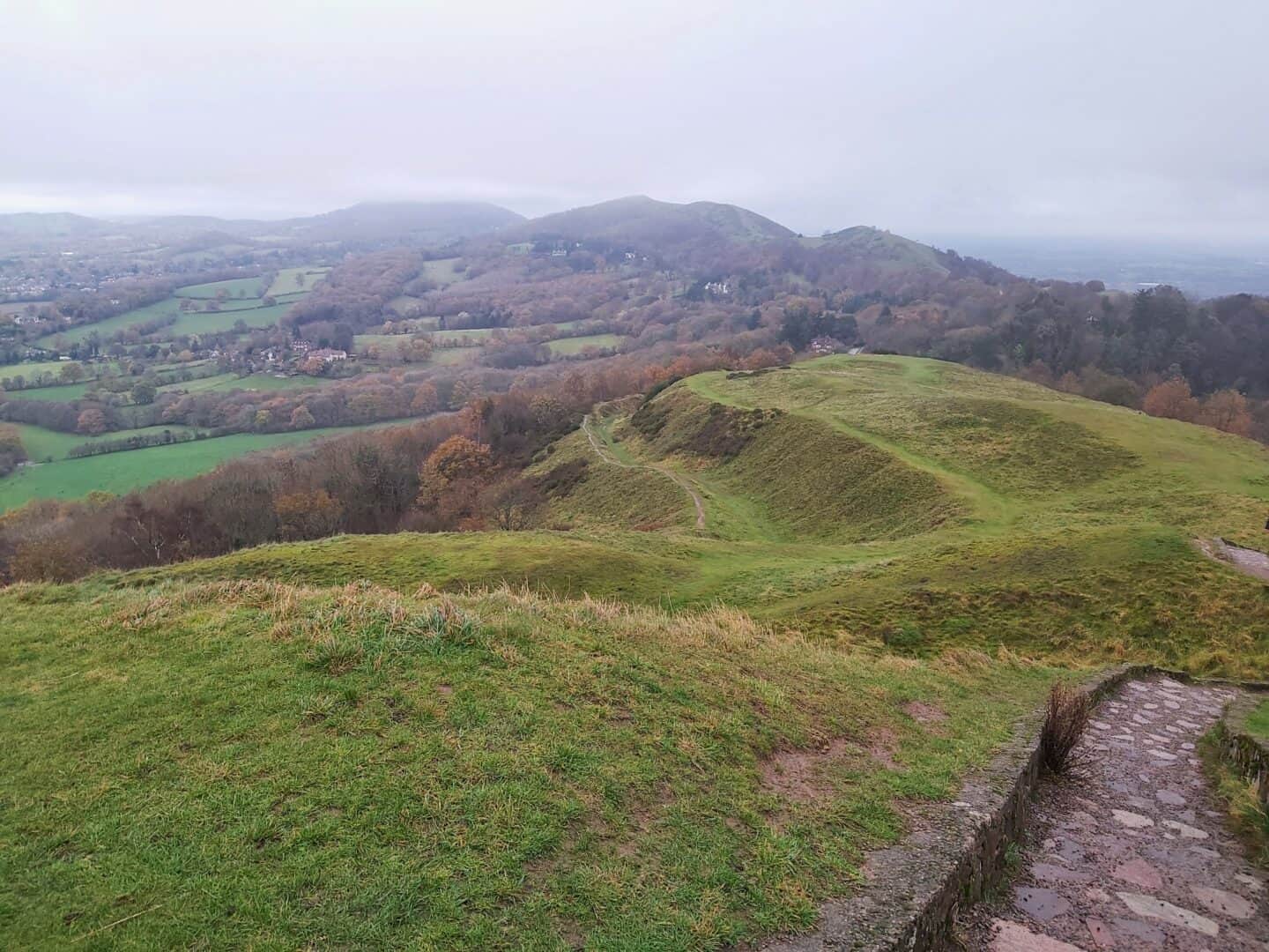Path on right hand side leading down grassy hillside with Malvern Hills stretching from point of photo to the horizon