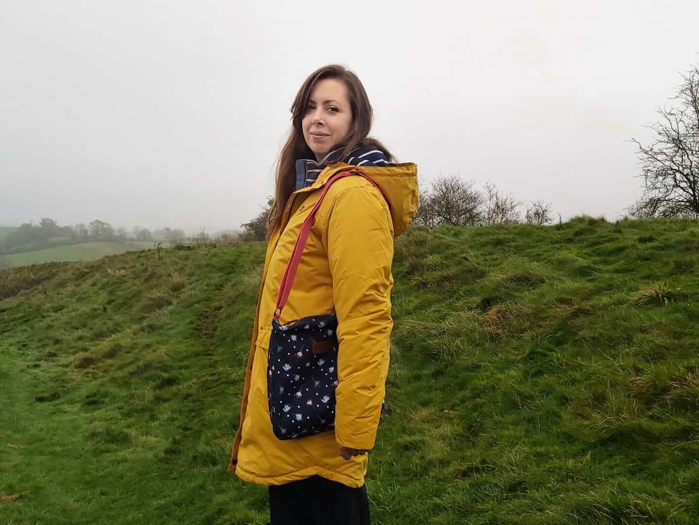 Wearing a yellow raincoat and blue bag with pink strap standing on grass on foggy day