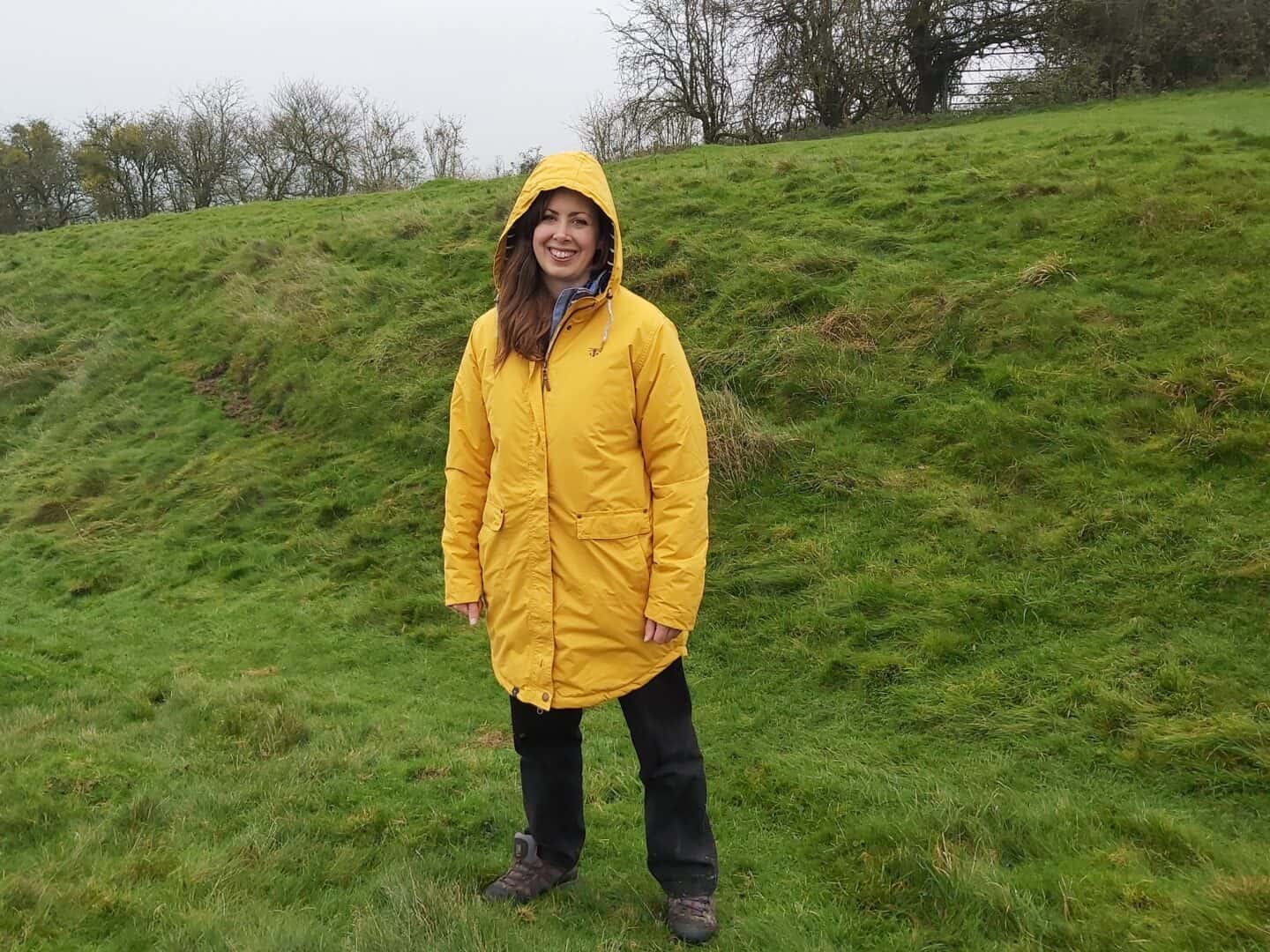 Facing towards the camera wearing yellow raincoat and black trousers with muddy shoes standing on grassy bank 