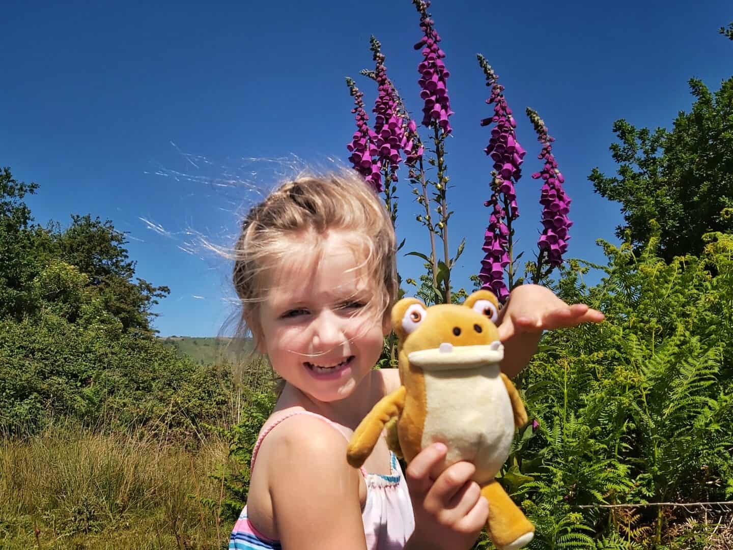 Birthday gifts by age: Girl showing off her cuddly dinosaur toy with foxgloves and blue sky in the background