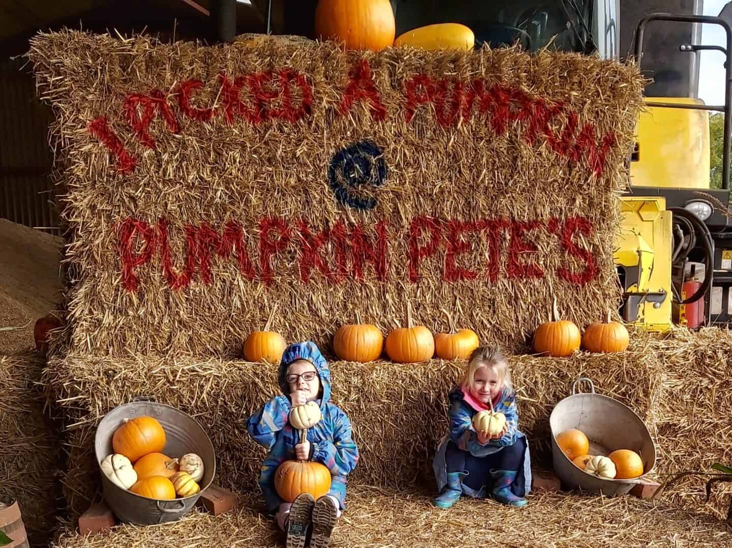 Two girls sitting on hay bales with Pumpkin Pete's written on the hay bales and pumpkins on top of them
