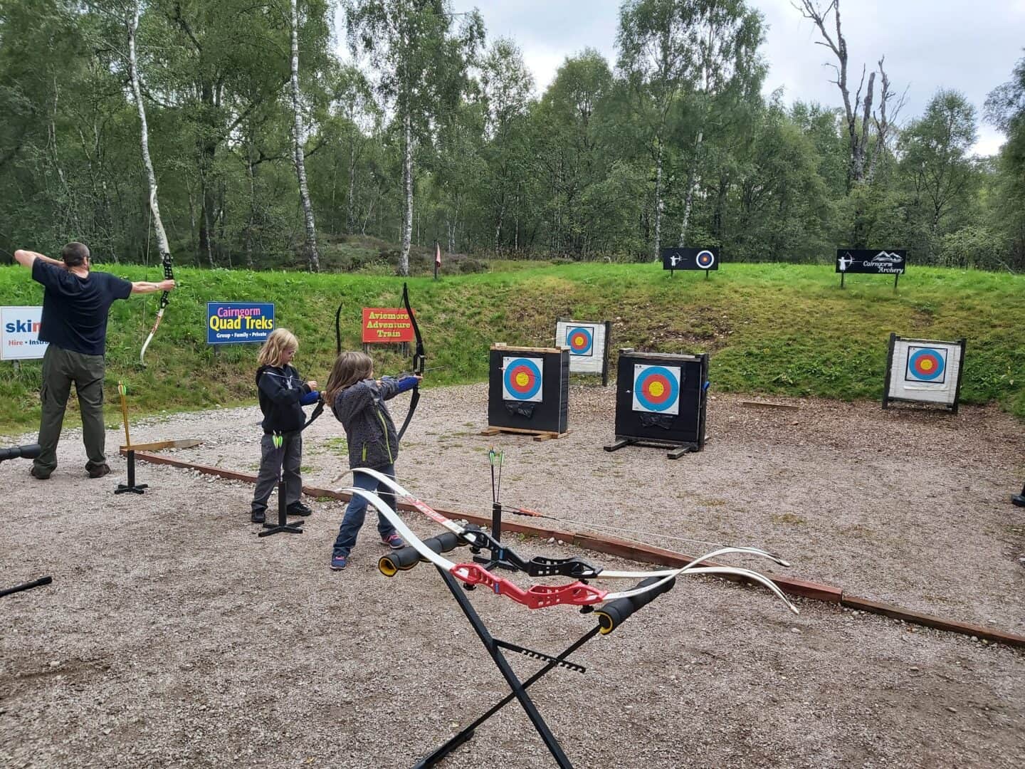 Two girls and a man doing archery at Dalraddy Holiday Park. Aiming their bows at targets with grassy bank and trees in background.  