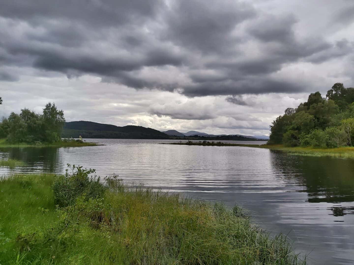 Loch Insh with grasses in foreground and Cairngorm mountain range in background on a cloudy day.  