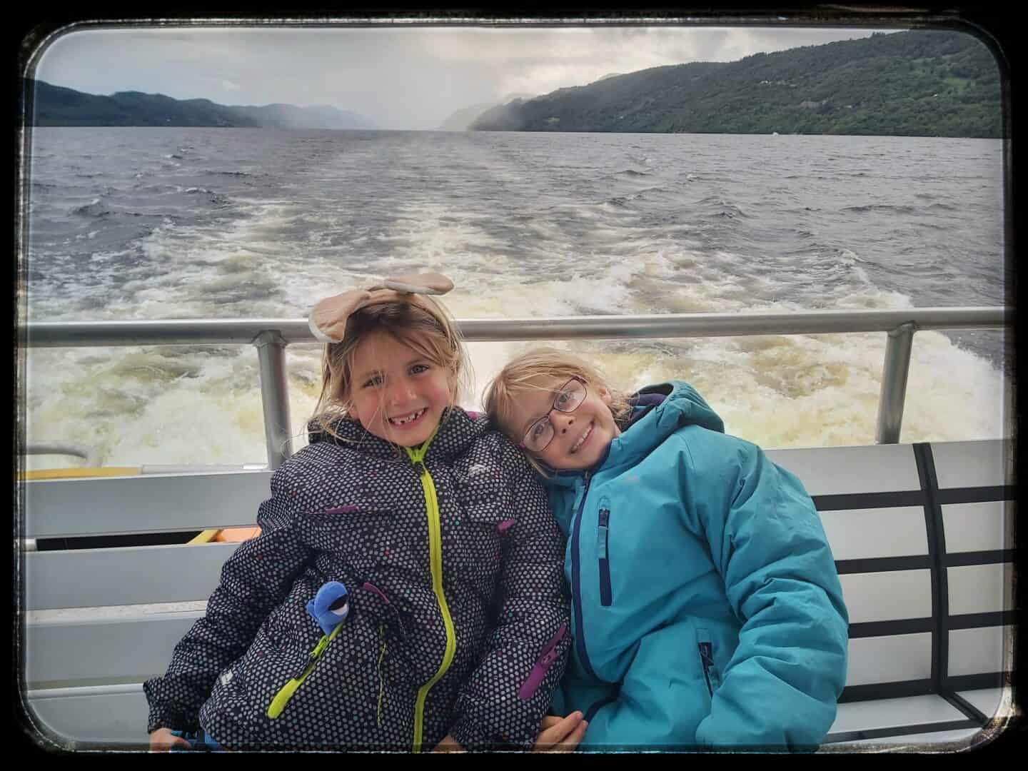 Two girls sitting on a bench on a boat in Loch Ness with water and mountains in background