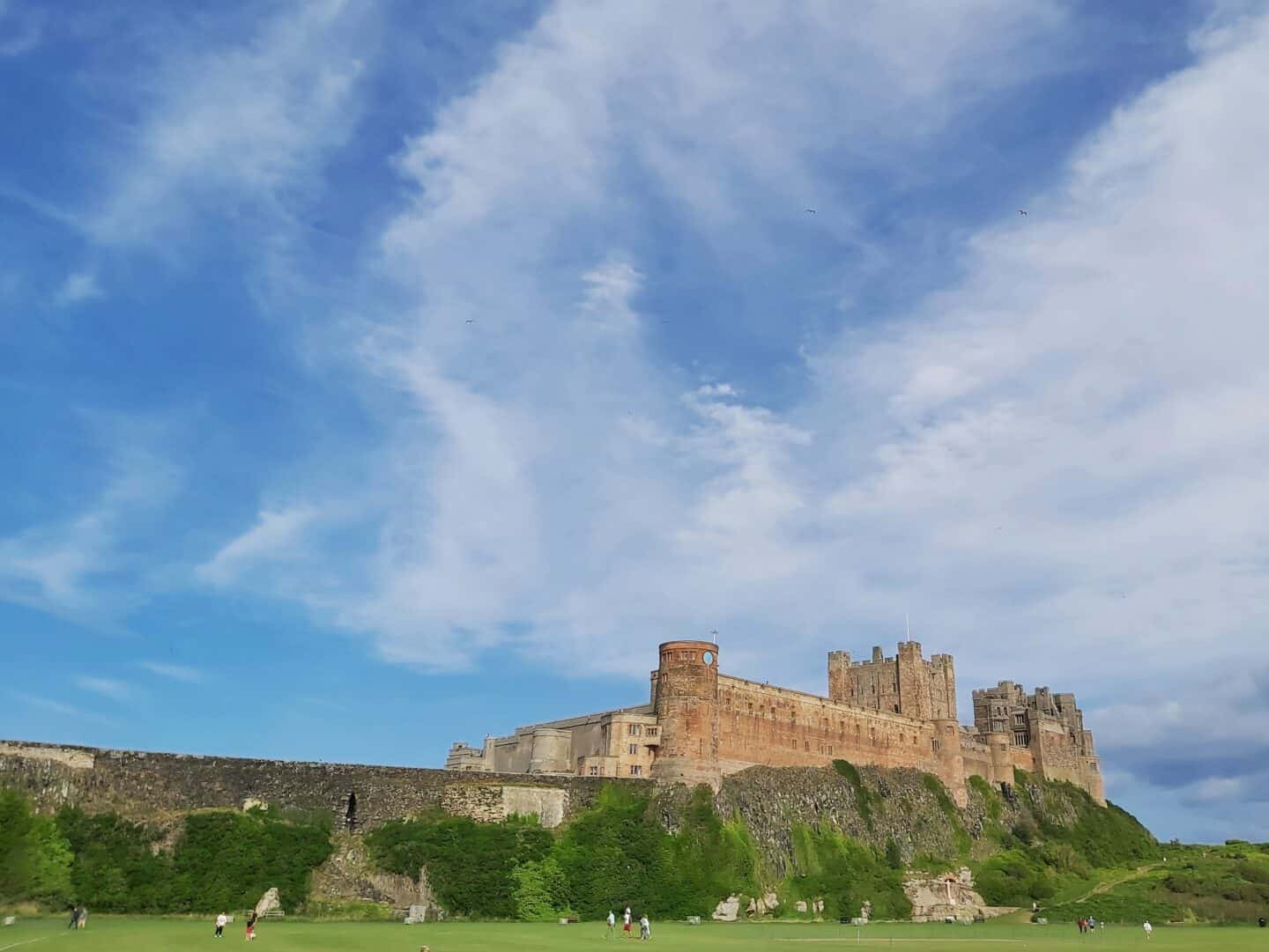 Family road trip itinerary: From the West Midlands to the Scottish Highlands. Spending a couple of days in Bamburgh. Photo of Bamburgh Castle with sports pitch in foreground and blue sky with light white clouds.  