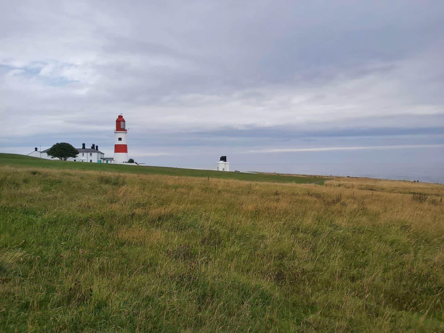 Family road trip itinerary: From the West Midlands to the Scottish Highlands. Breaking the journey from Bamburgh to Yorkshire at Souter Lighthouse. Photo of the lighthouse, taken from grassy bank with lighthouse in middle distance. Lighthouse is traditional white with red stripe and red top.  