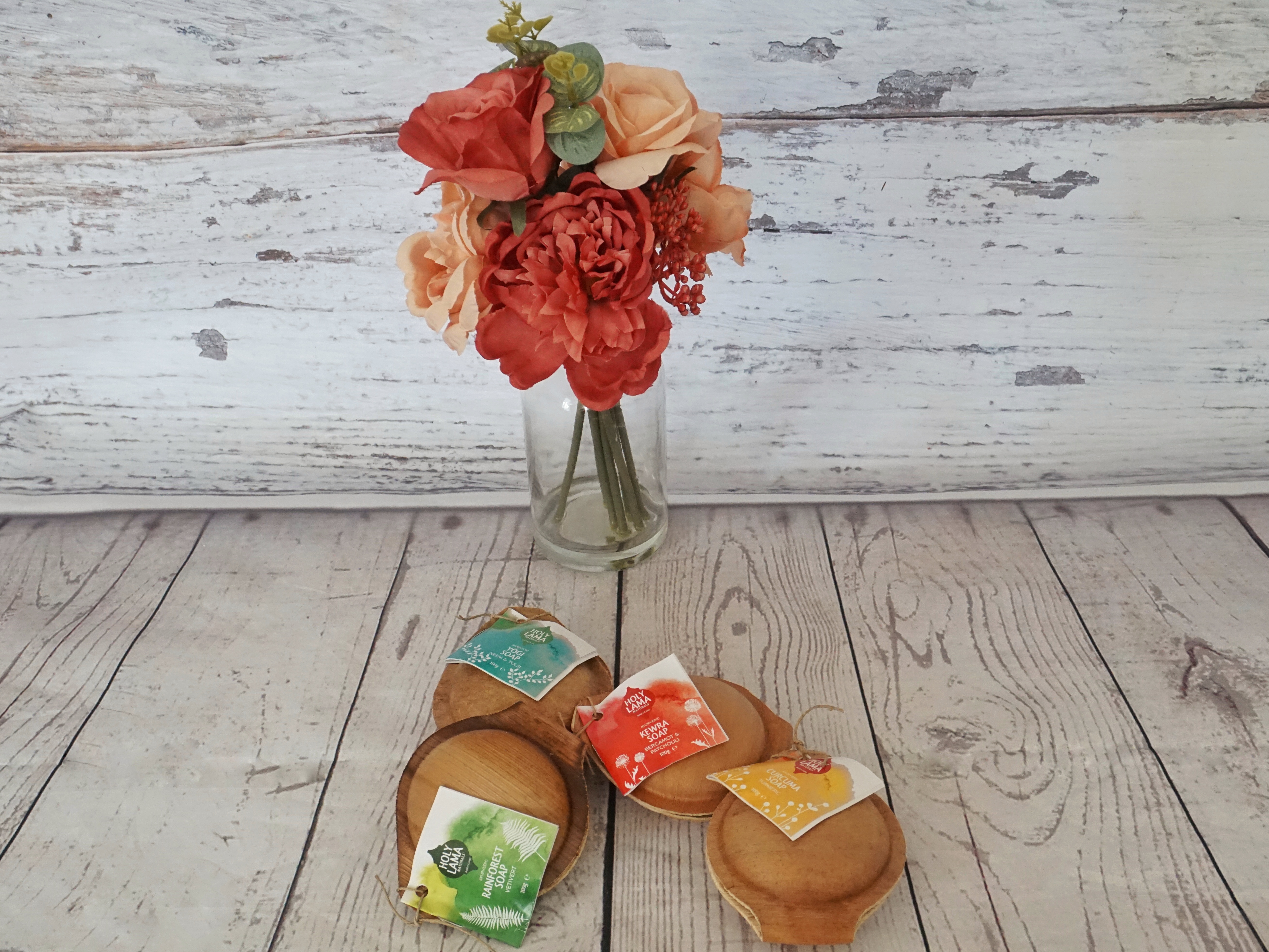 Four Holy Lama Soaps presented against a wooden background with red and peach flowers in a vase behind