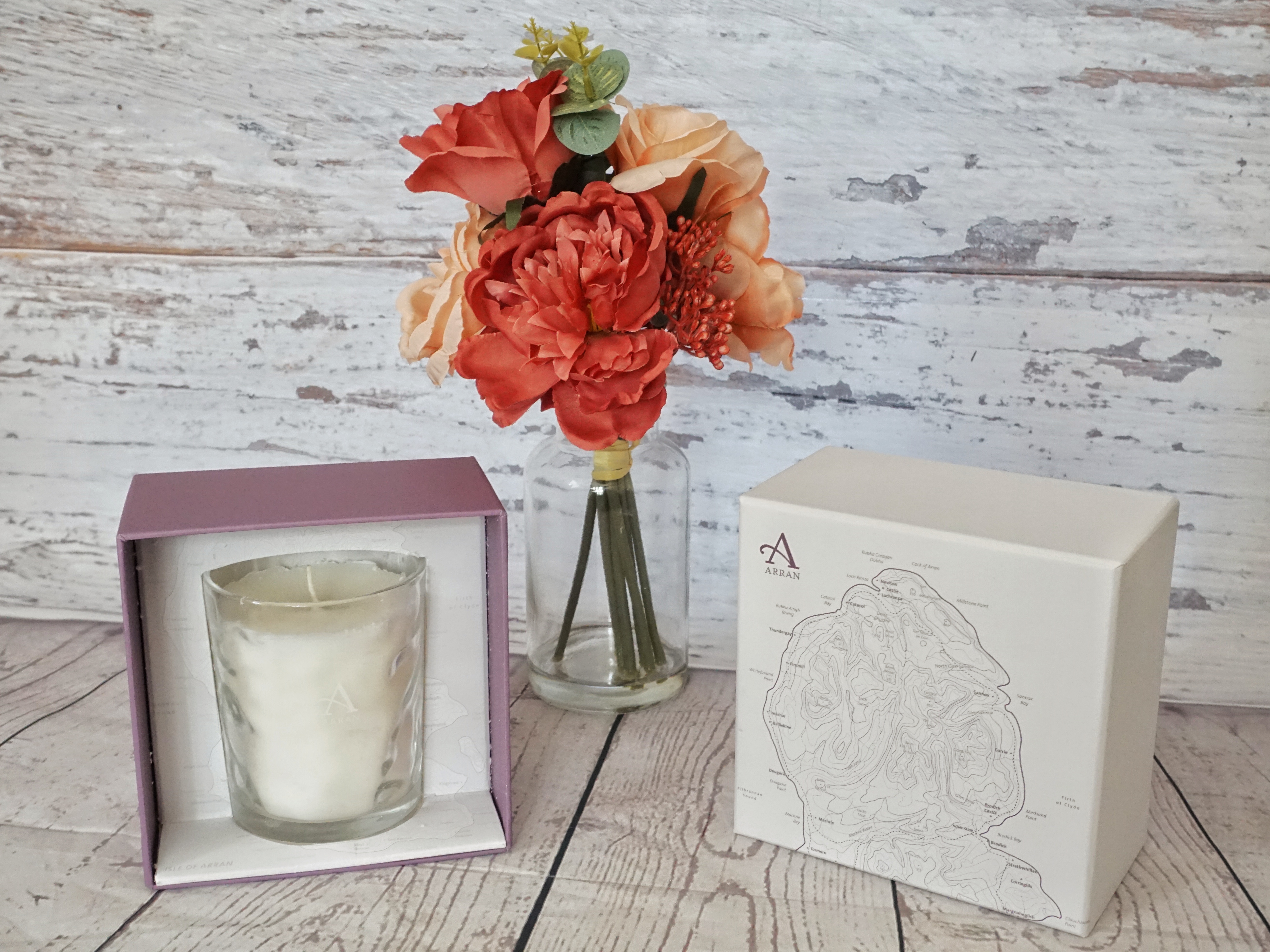Arran Sense of Scotland Candle presented against a wooden background with red and peach flowers in a vase behind