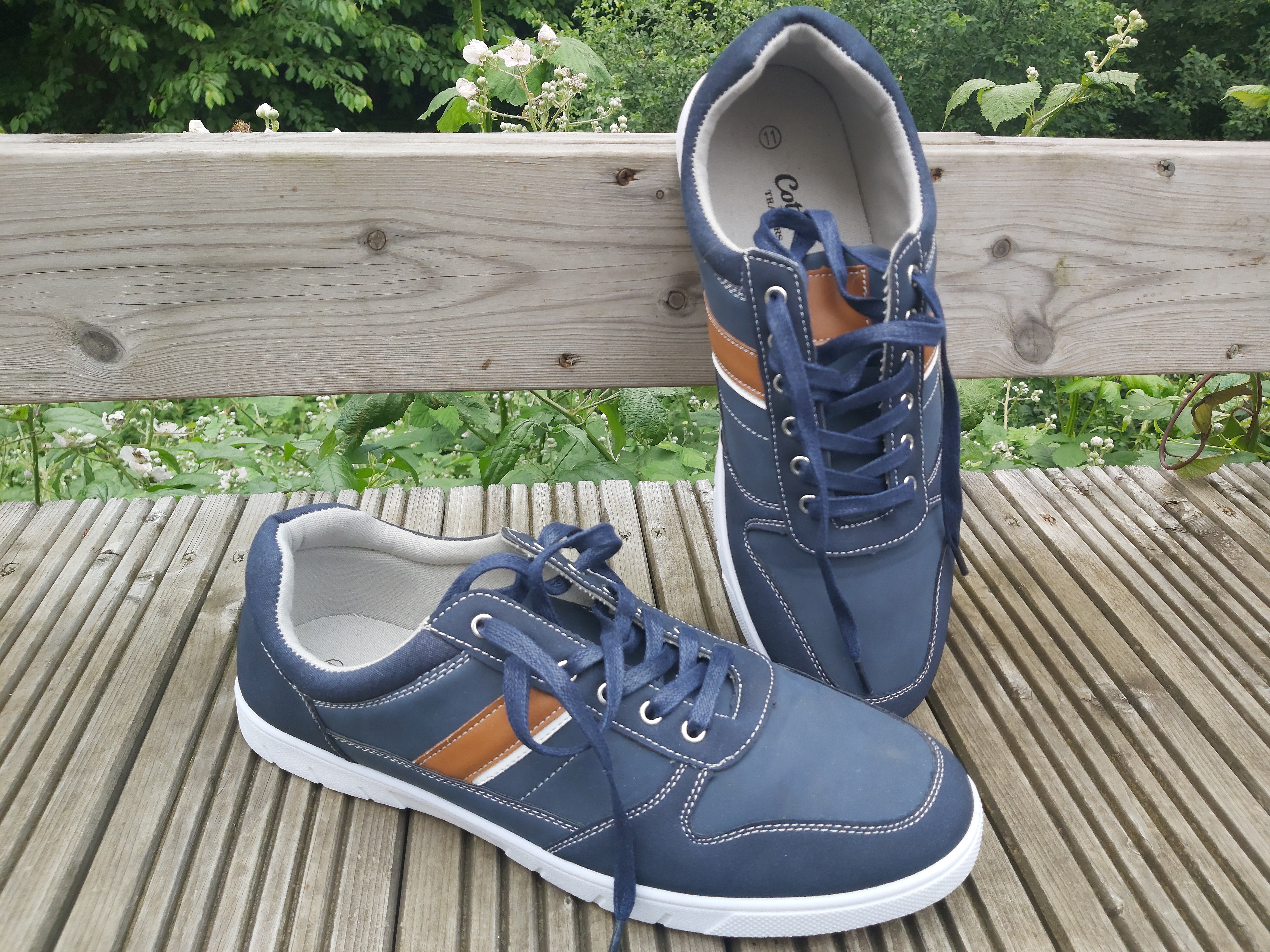 Casual lace up trainers in blue and orange with white stripe and blue laces displayed on wooden background with greenery behind.  