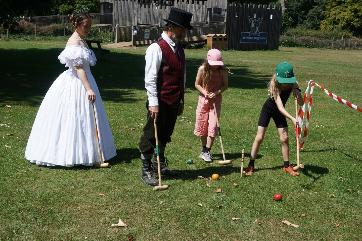 Children playing croquet with Victorian characters on a grass lawn