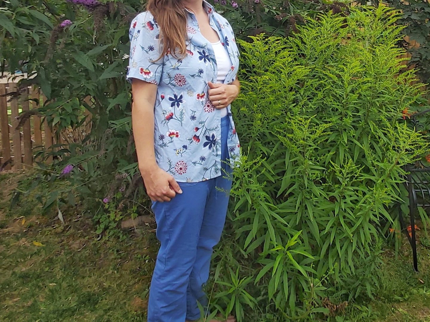Adult female pictured from the neck down wearing shirt and bright blue trousers standing beside a green bush