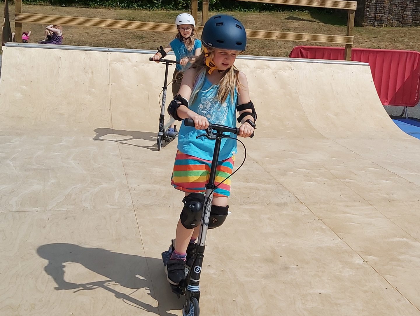 Two girls on scooters on a skate ramp with helmets and knee pads on