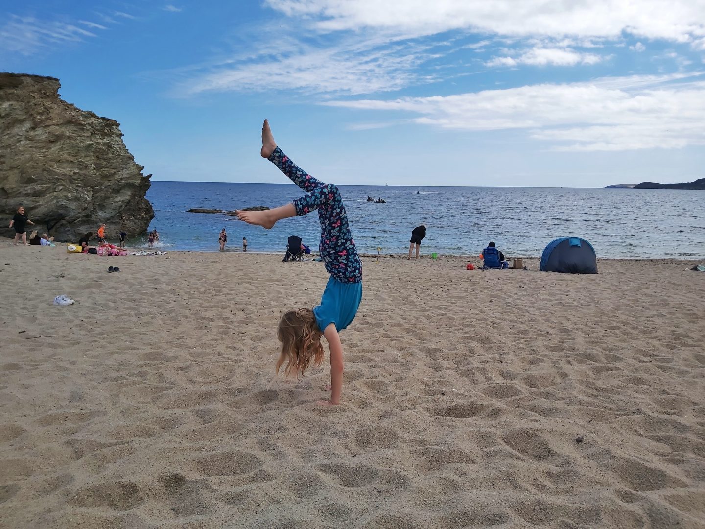 Girl doing handstand on a sandy beach with blue sky and sea behind and a large rock to left hand side