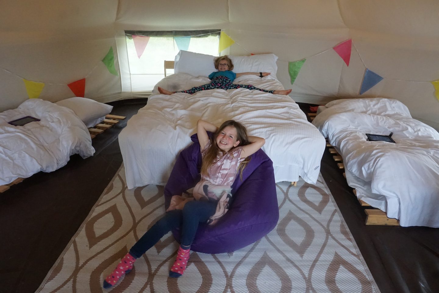Inside of a bell tent at YHA Eden Project campsite with double bed and two single beds. Girl lying on double bed and another girl sitting on a beanbag in front of it. 