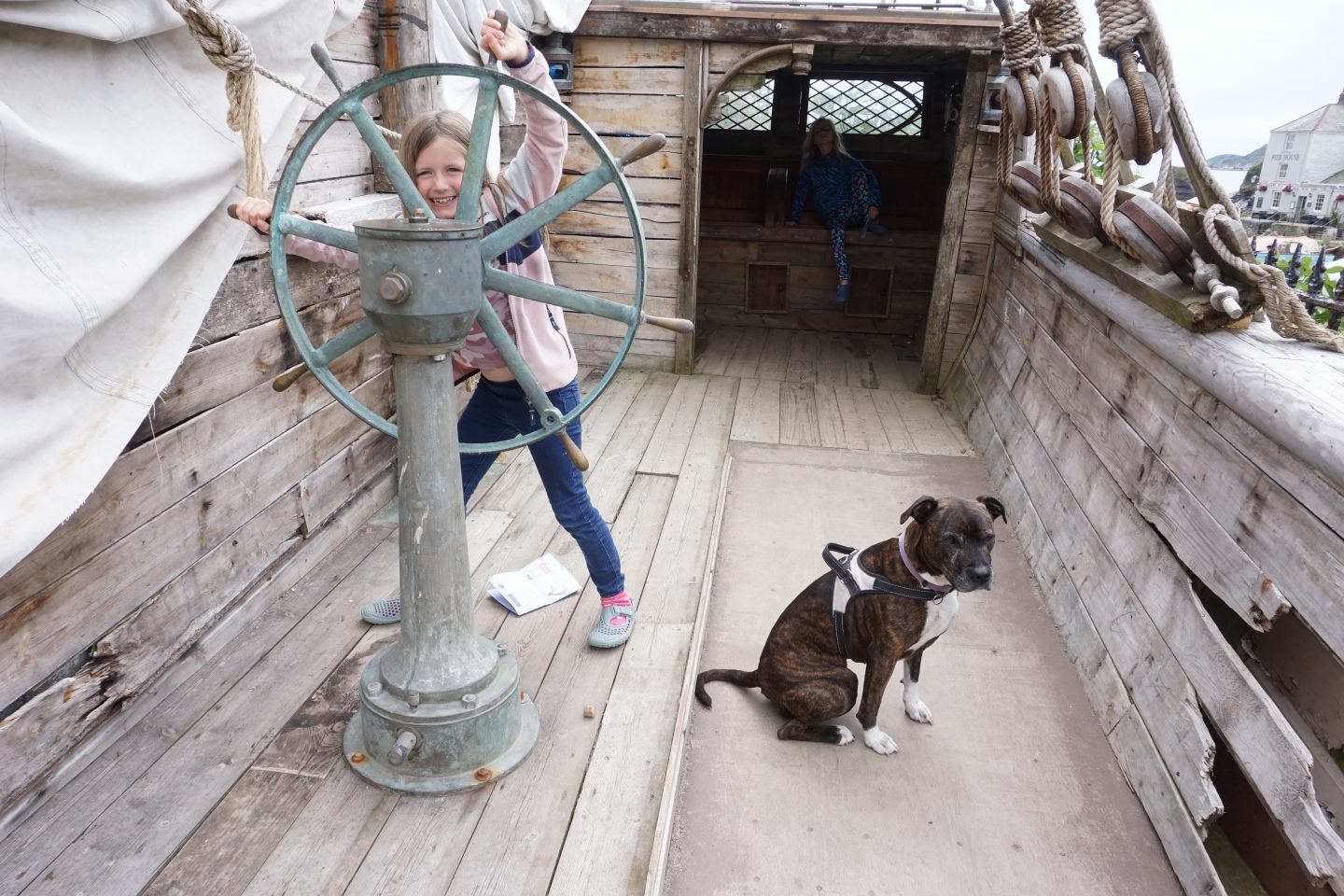 Girl turning a ship steering wheel with brown staffie dog beside her and girl behind in cabin. Shipwreck Treasure Museum in Charlestown