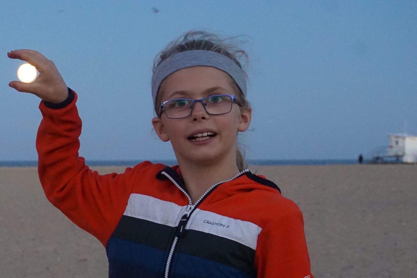 Girl wearing glasses and a grey hairband holding the moon on Great Yarmouth beach at dusk