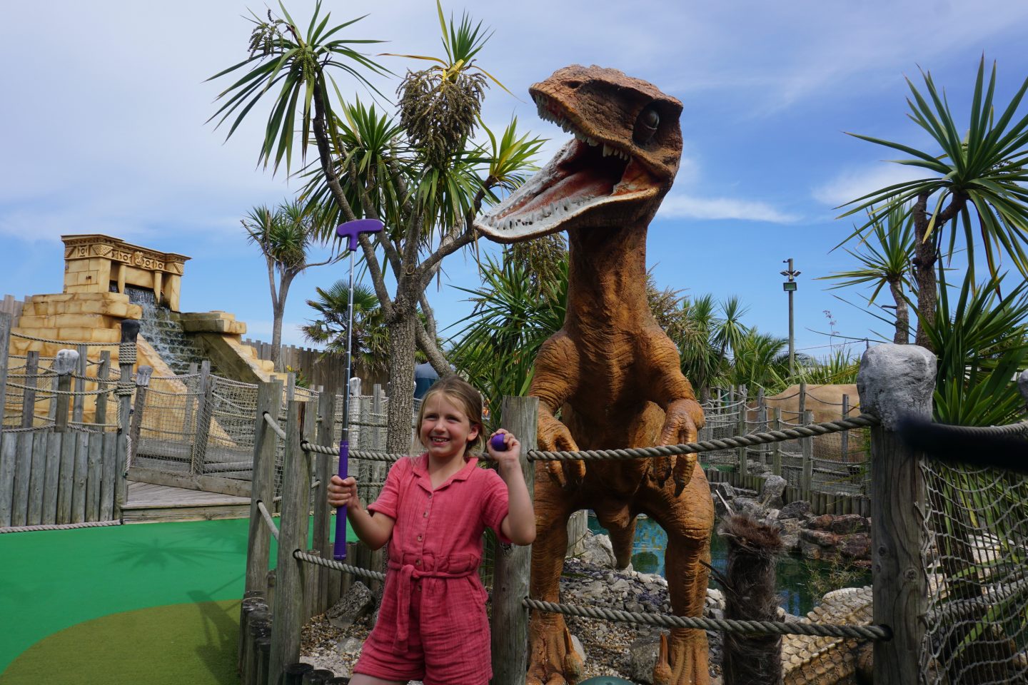 Large dinosaur model at Lost World Adventure Golf Great Yarmouth towers over little girl holding a golf club and golf ball with palm trees in the background