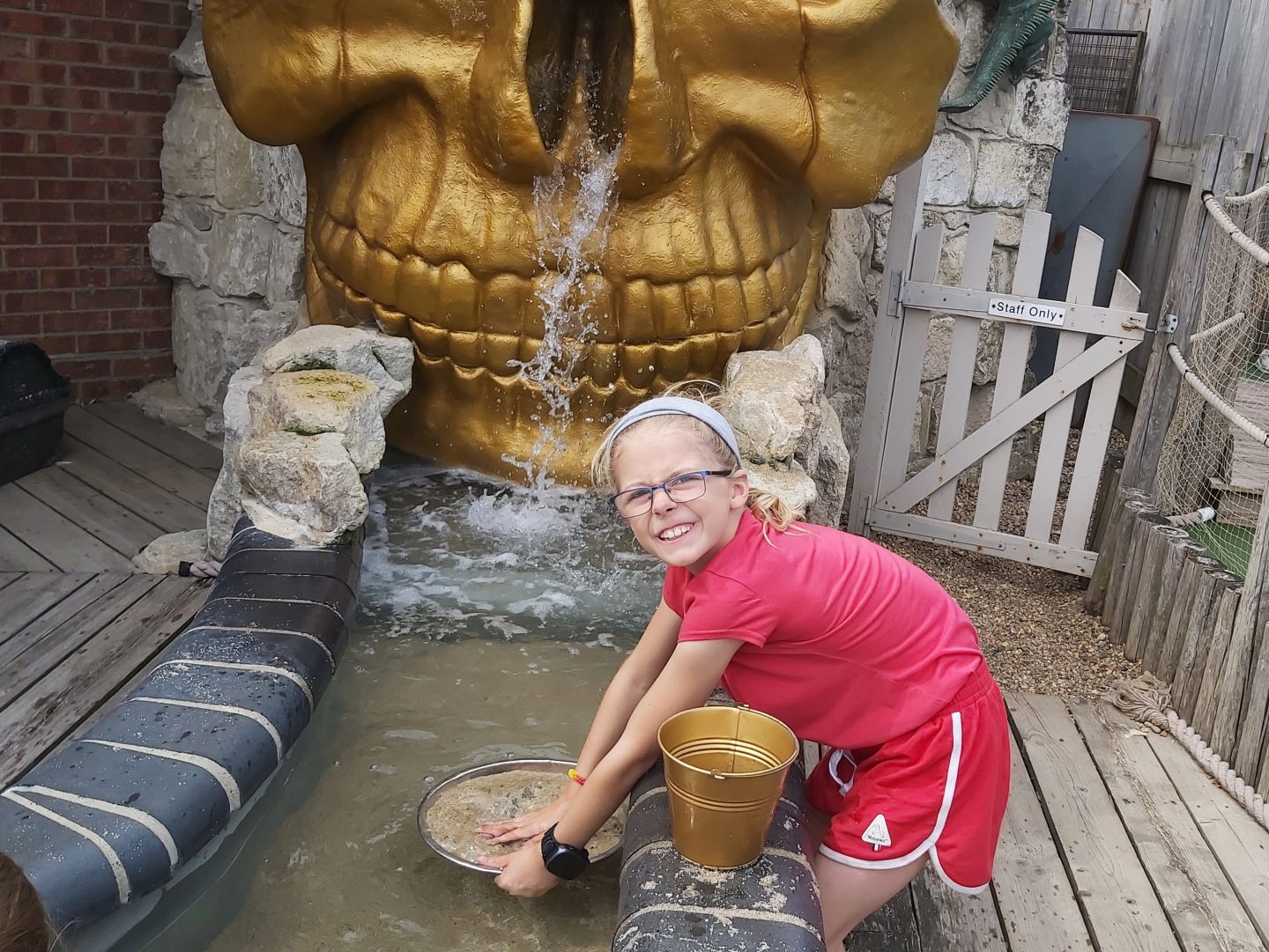 Girl in red t-shirt and shorts panning for gold in a stream outdoors in front of a large gold skull with a gold bucket beside her at Lost World Adventure Golf Great Yarmouth