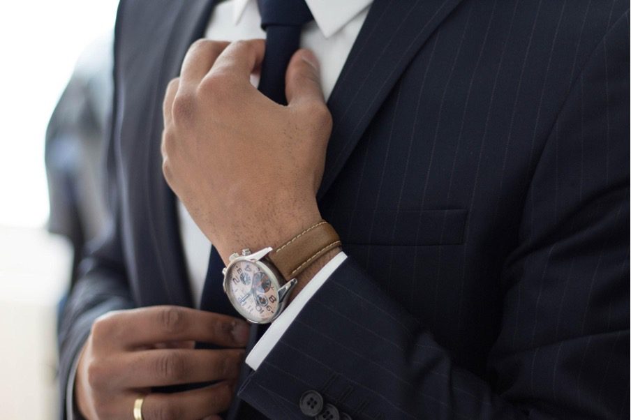 Man wearing black suit and tie and white shirt with brown watch strap on silver faced watch