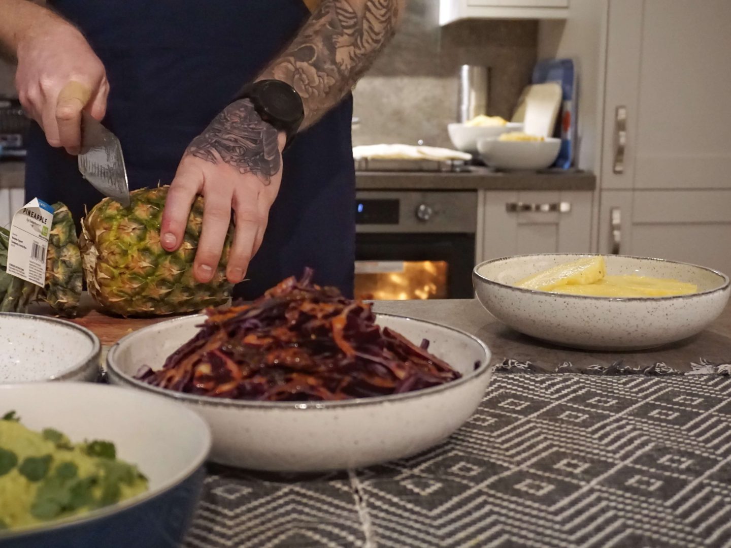 Hands of chef Dean with tattoos and a watch on left wrist he is cutting a fresh pineapple with spiced cabbage in front of him and other bowls of mexican food around