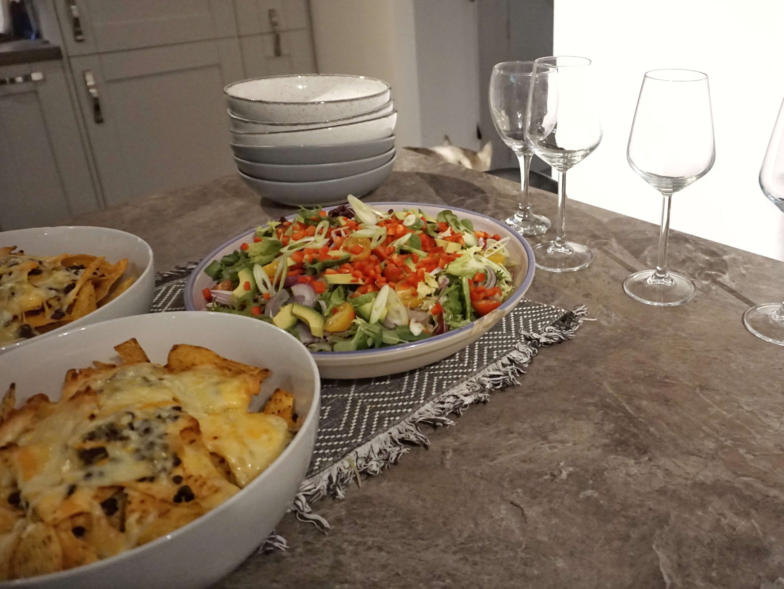 Fresh salad beside wine glasses and bowls stacked up for serving with loaded nachos in the foreground