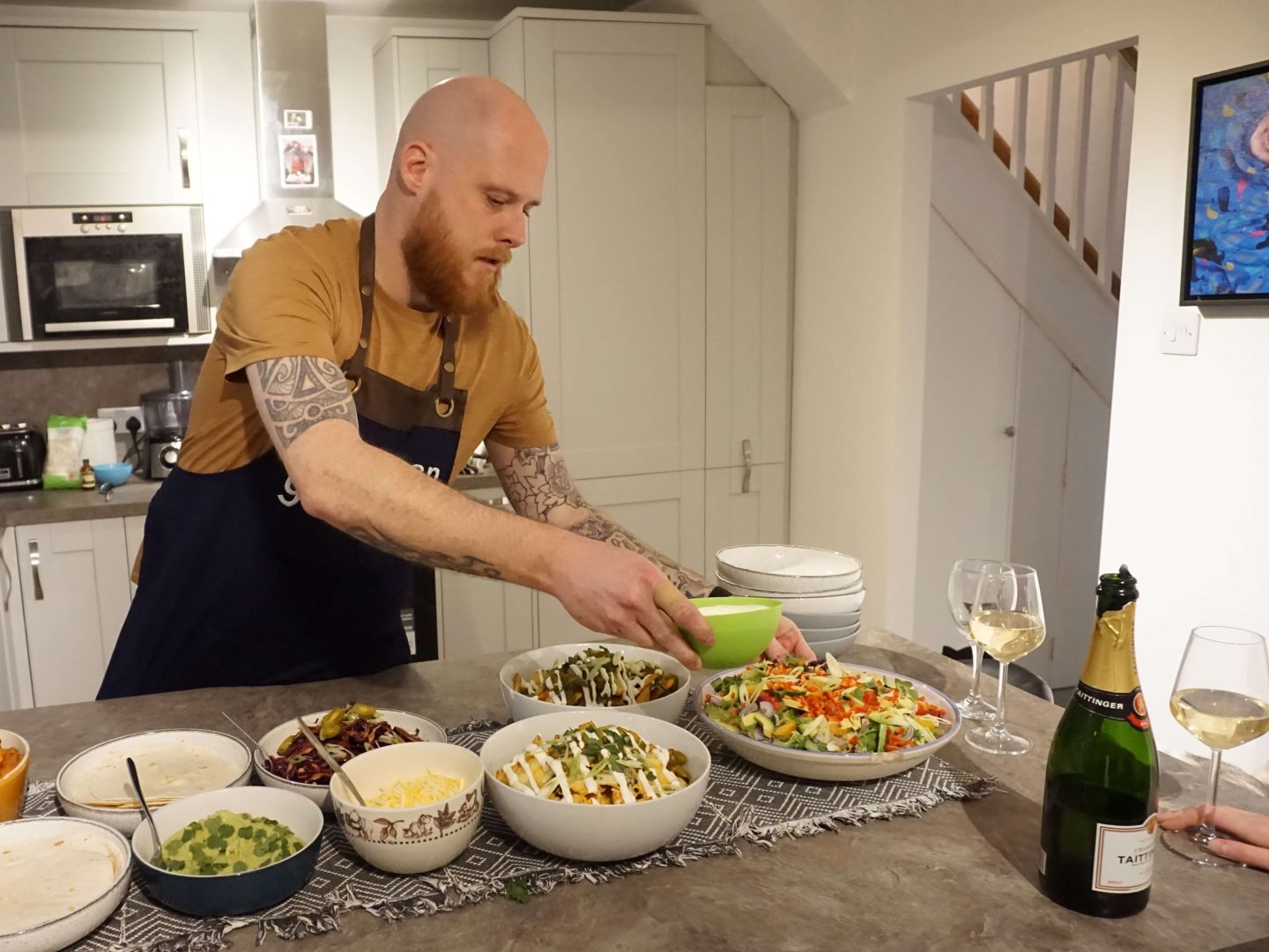 Chef Dean from yhangry putting out bowls of mexican food in a white kitchen with a bottle of champagne on the table in the foreground 
