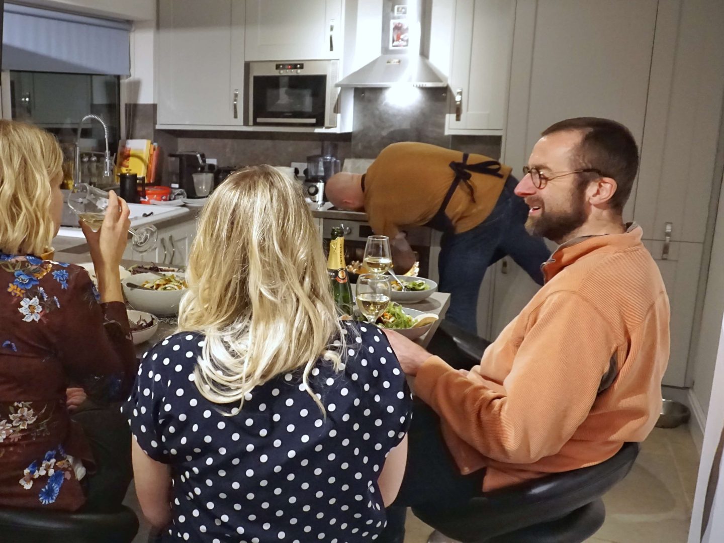 My husband and two sisters sitting at a table in the kitchen with bowls of Mexican food and wine on the table and yhangry chef Dean in the background putting something in the oven