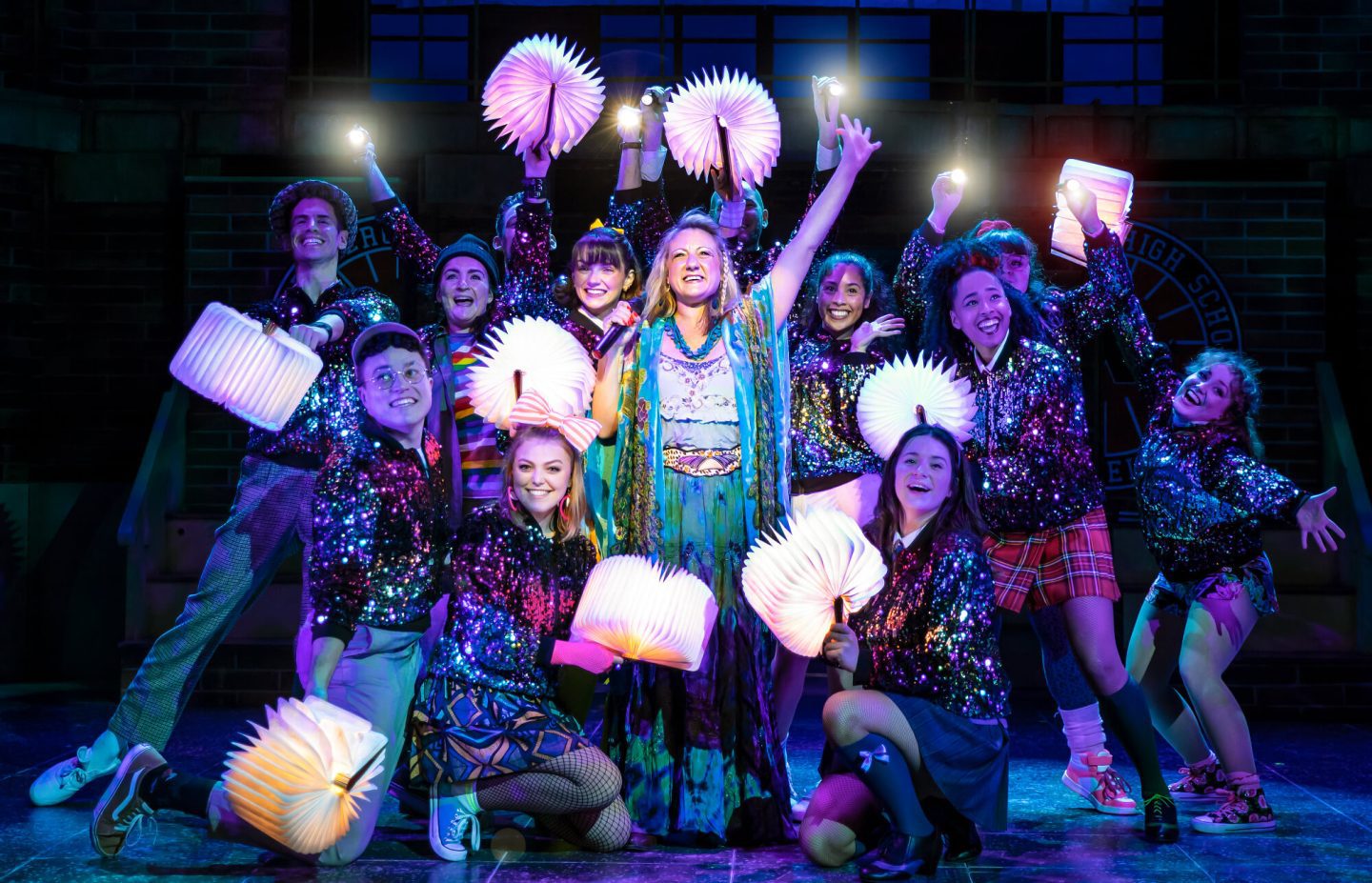 Heathers the Musical cast holding fans that light up and standing all together, some also holding torches.