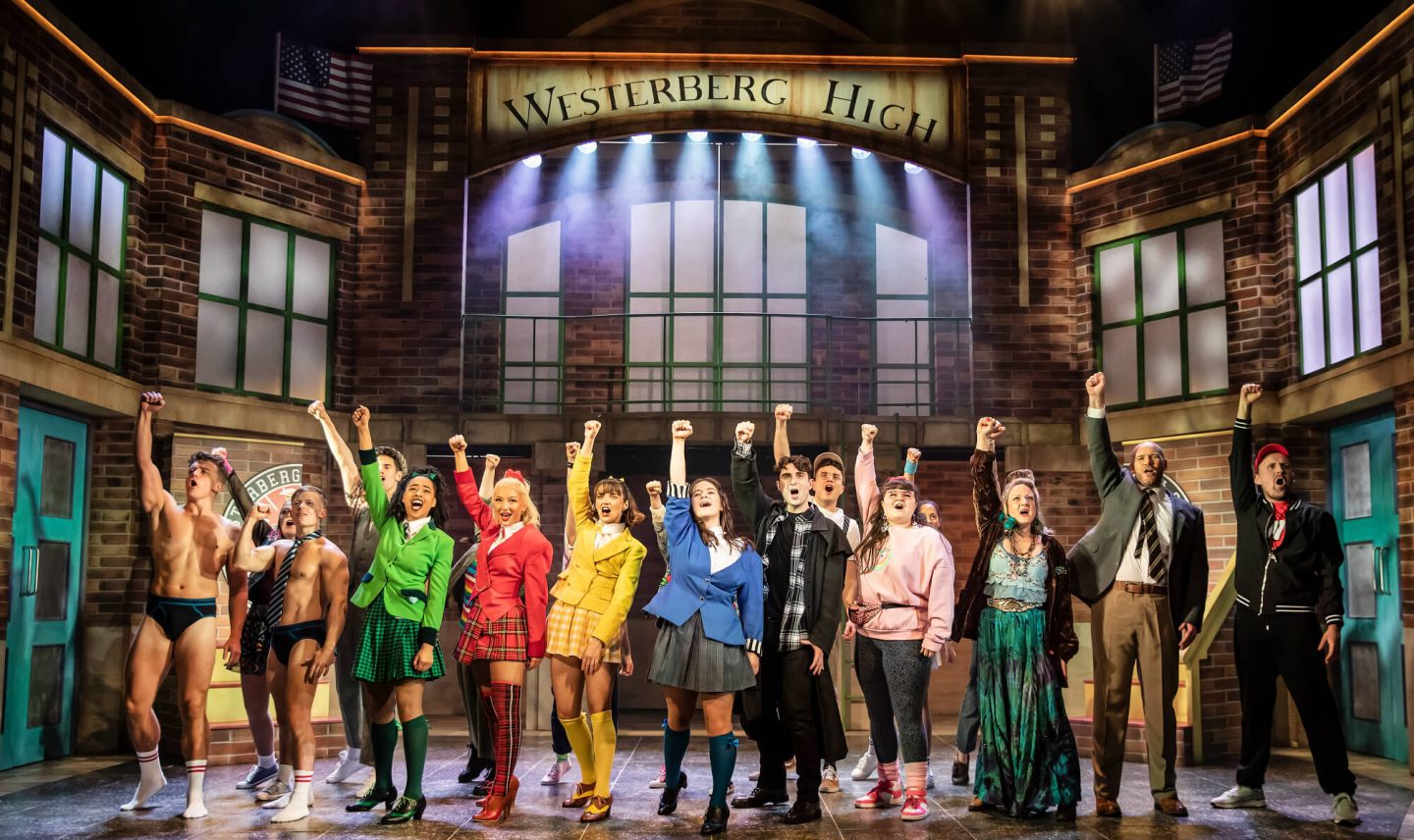 Heathers the Musical cast on stage at the end of a song, all raising their right arms in the air.
