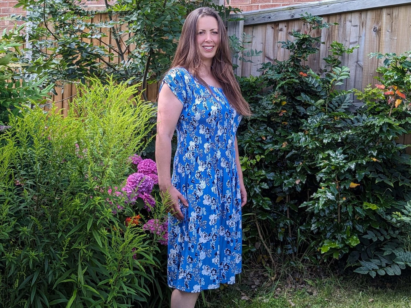 Natalie standing in the garden wearing a blue floral Cotton Traders Jasmine Harman Collection dress