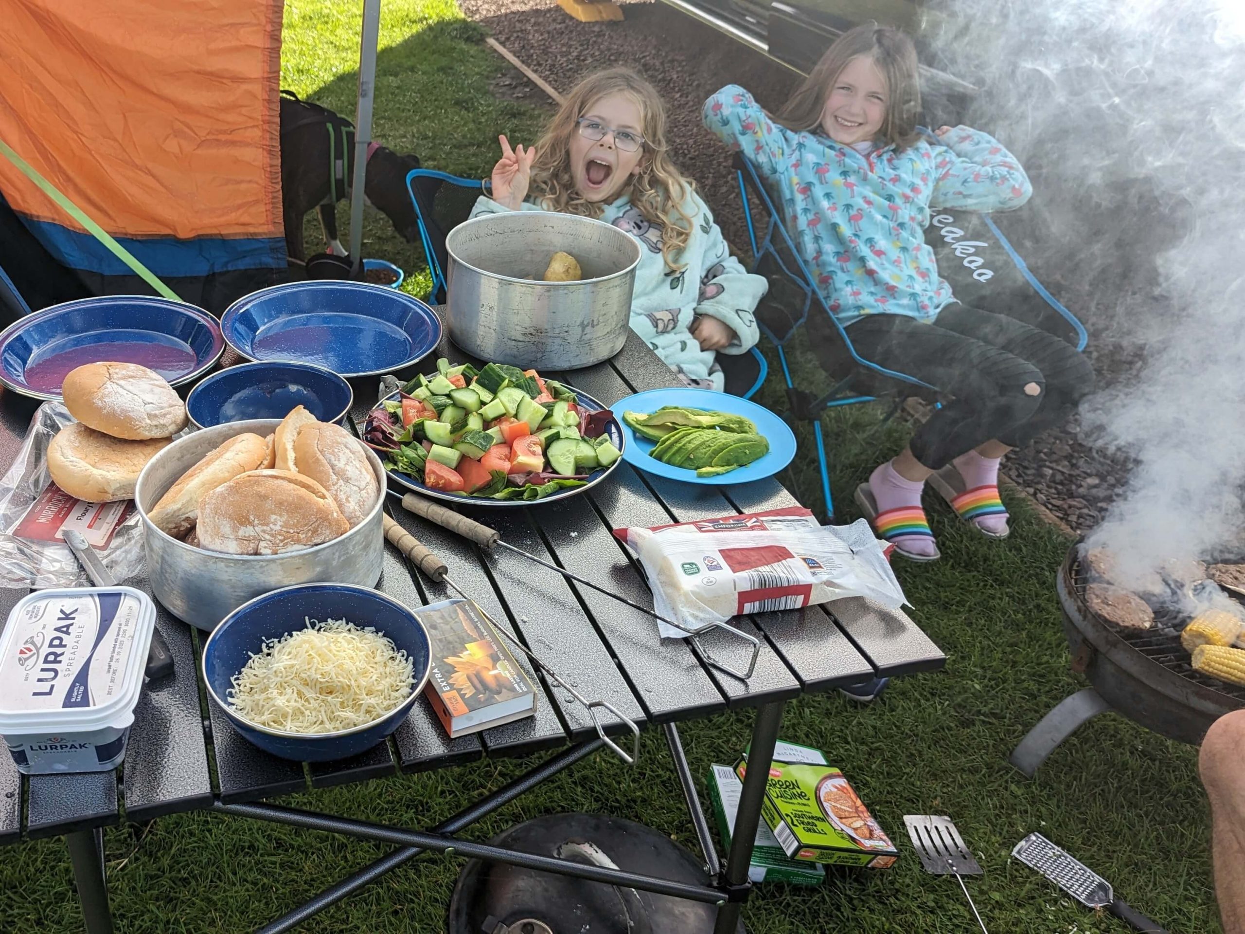 Two girls smiling sitting by a folding camping table with food on and BBQ smoke to left hand side of image