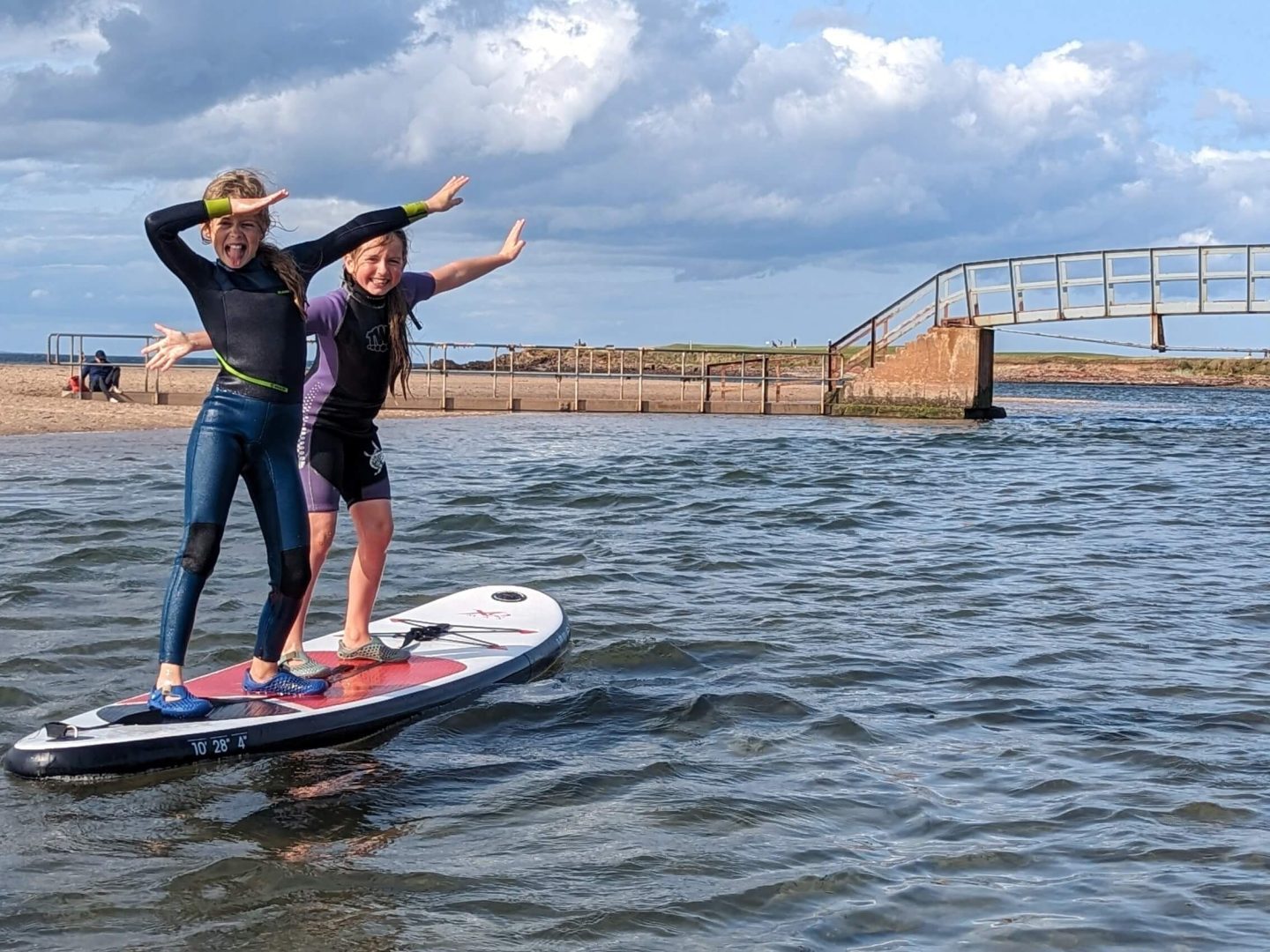Two girls wearing wetsuits dancing on a paddleboard on the sea with a bridge and beach in the background