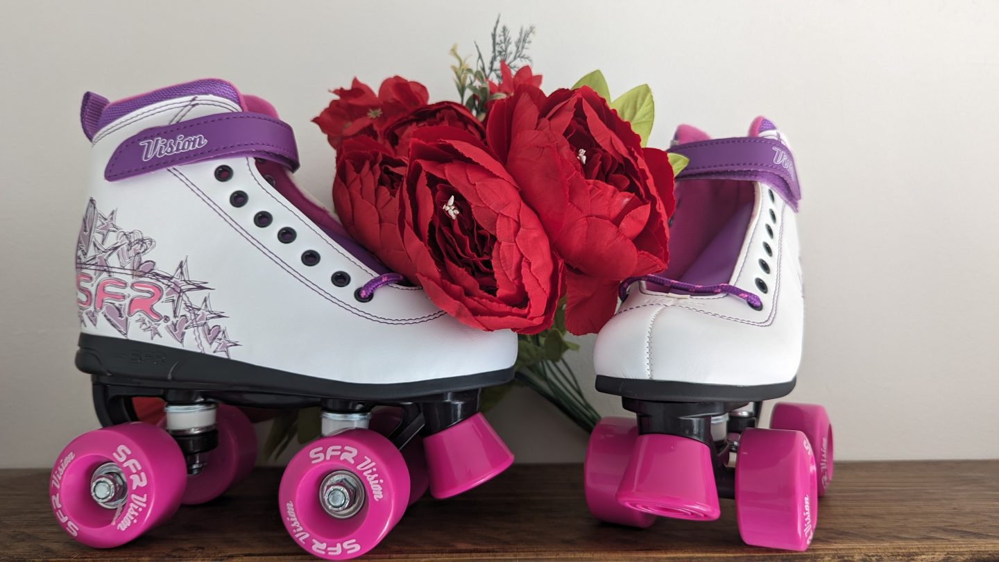 White roller skates with purple wheels and detailing displayed on a wooden shelf with red flowers behind