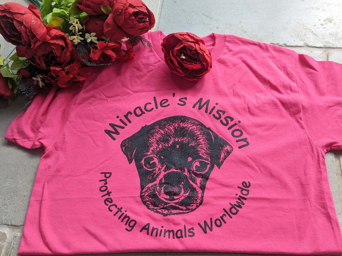 Miracles mission charity t-shirt in bright pink with a picture of a dog on the front displayed beside red flowers on a tiled background