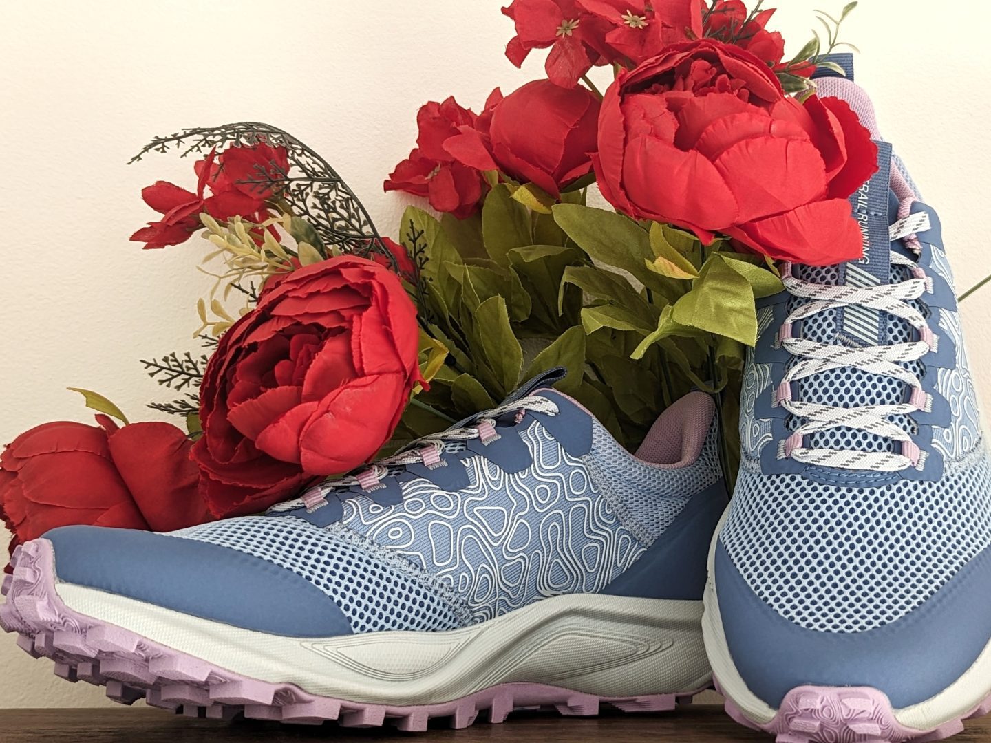 Light blue Helly Hansen Featherswift Trail Runnings Shoes pictured against a white wall with red flowers behind