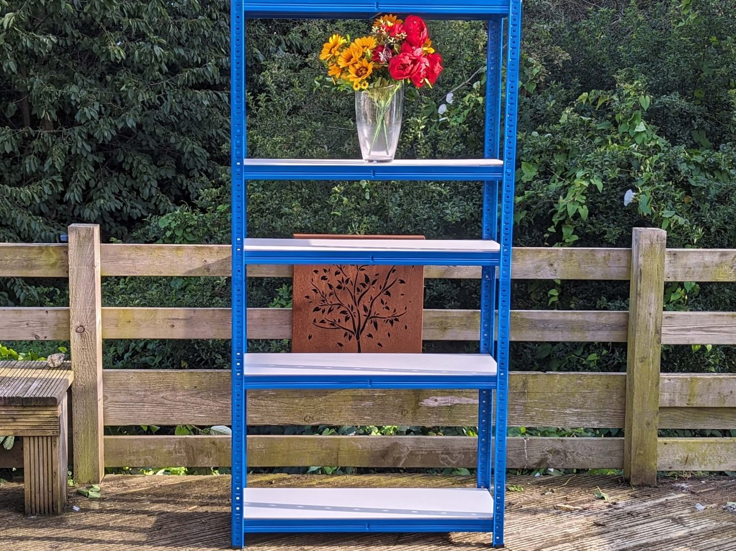 Blue-edged shelving unit displayed on a wooden decking with a vase of red and yellow flowers on the top shelf