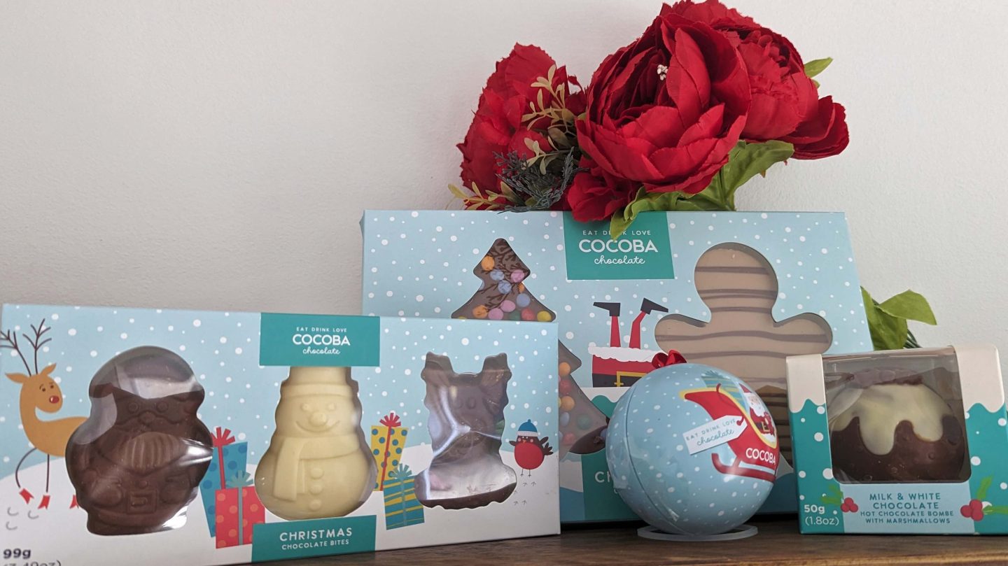 Cocoba Chocolate and Hot Chocolate Christmas Gifts displayed on a wooden shelf with red flowers behind