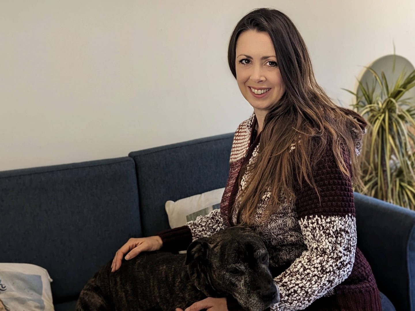 Natalie sitting on a blue sofa next to Bubbles, a dark brown brindle staffie cross dog. Natalie is wearing a zip up hoodie style cardigan in red and beige from Cotton Traders Autumn Winter knitwear collection. 