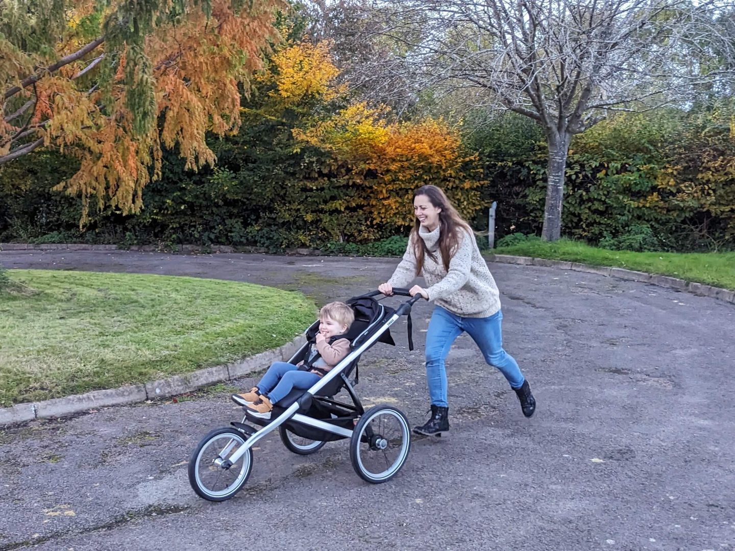 Natalie running around a road roundabout pushing a running buggy with her nephew in, both are smiling and trees in the background have red and orange autumn leaves