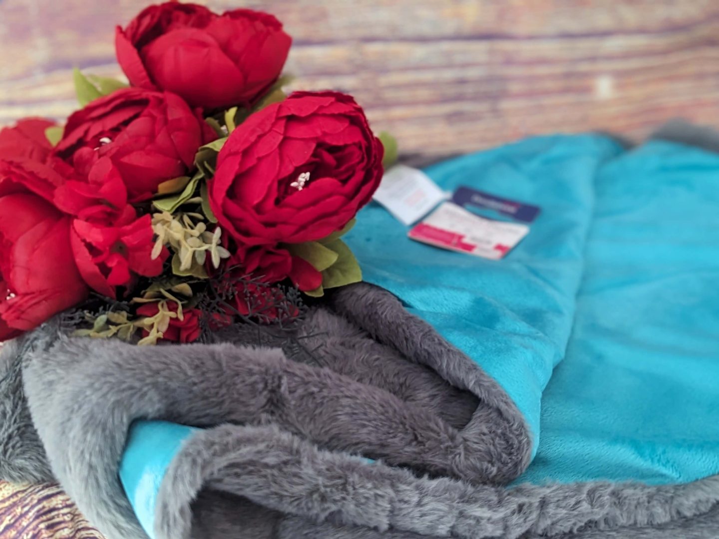 Turquoise and grey doodlebone dog blanket displayed against a wooden backdrop with red flowers beside