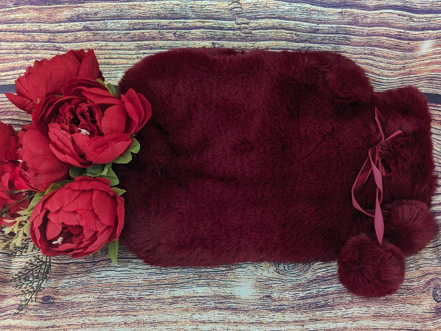 Red faux fur hot water bottle from Vendula London displayed against a wooden backdrop with red flowers beside